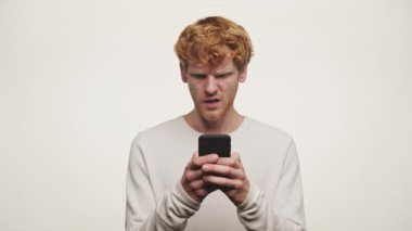 Negatif ifadeli kızıl adam, beyaz arka planda izole edilmiş akıllı telefon kullanarak kötü haberleri okuyor.