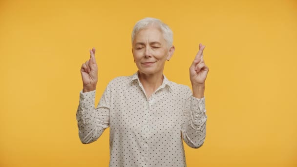 目を閉じたシニア女性が指を交差し 黄色の背景に希望する — ストック動画