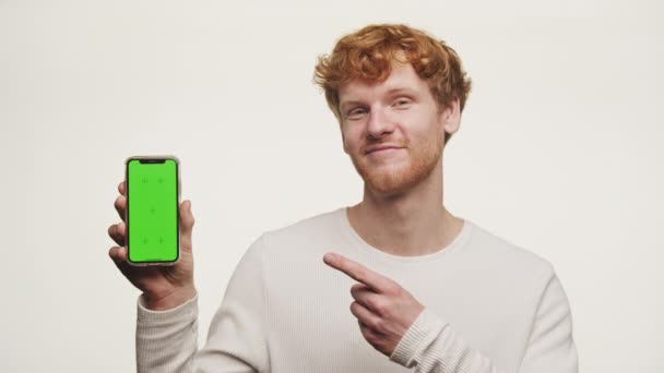 笑着的红头发男人指着智能手机上的绿色屏幕 白色背景隔离起来 — 图库视频影像
