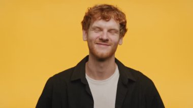 Neşeli kızıl saçlı adam, sarı arka planda izole edilmiş, dostane bir gülümsemeyle, günlük giysiler içinde başını sallıyor.