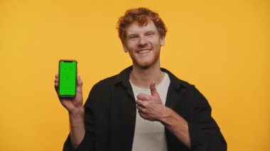 Neşeli adam, sarı arka planda izole edilmiş yeşil ekranlı akıllı telefonu sunarken başparmağını kaldırıyor.