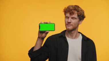 Kendine güvenen genç adam elinde yeşil ekranlı bir akıllı telefon tutuyor, onaylama jesti ile başını sallıyor, uygulama ekranına hazır, sarı arka planda izole edilmiş.