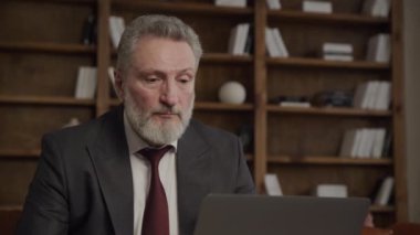 Olgun iş adamı, modern bir çatı katındaki masasında bilgisayarını kullanarak görüntülü konuşma yapıyor.