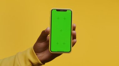 Yeşil ekranlı akıllı telefonu tutan bir elin yakın çekimi, sarı bir arkaplan üzerine kurulmuş.