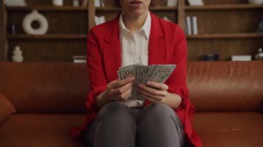 Şık kırmızı ceketli bir kadının yüz dolarlık banknotları, finans ve başarı kavramlarını sayarken yakın plan fotoğrafı.