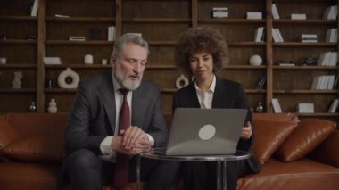 Daha yaşlı bir erkek yönetici ve genç bir kadın profesyonel video görüşmesi. Sofistike bir ofis ortamında dizüstü bilgisayar kullanarak etkili bir çapraz-kuşak işbirliği gösteriyorlar.