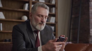 Takım elbiseli olgun bir işadamı akıllı telefonuna odaklanmış, modern bir ofis ortamında oturuyor.