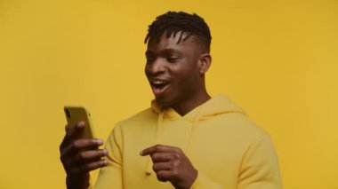 Akıllı telefonuyla gülümseyen neşeli Afro-Amerikan adam sarı kapüşonlu sarı kapüşonlu güzel haberi kutluyor.