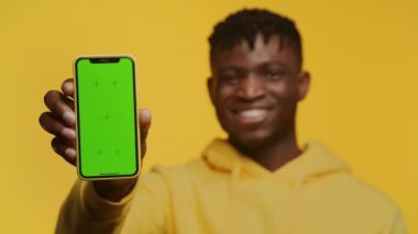 Neşeli Afrikalı Amerikalı adam yeşil ekranlı cep telefonu sergiliyor. Uygulamaya hazır, sarı arka planda izole edilmiş.