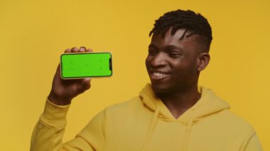 Sarı kapüşonlu gülümseyen Afrikalı erkek yeşil ekranlı akıllı telefonu sunuyor. Uygulama modelleri için ideal, sarı arka planda izole edilmiş.