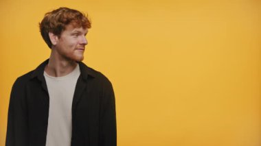 Siyah ceketli neşeli kızıl saçlı adam büyük bir gülümseme ile yan tarafı işaret ediyor, canlı sarı bir zemin üzerine kurulmuş