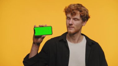 Kıvırcık kızıl saçlı genç adam yeşil ekranlı akıllı bir telefon tutuyor, parlak sarı bir arka plana karşı jest yapmadan başını sallıyor.