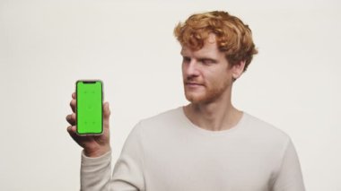 Genç kızıl saçlı bir adam elinde yeşil ekranlı bir akıllı telefon tutuyor. Beyaz arka planda izole edilmiş bir şekilde baş parmağıyla bir jest yapıyor.