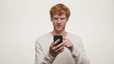 Kızıl saçlı genç adam, beyaz bir arka planda izole edilmiş akıllı telefonuna bakarken tiksindirici bir ifade takınıyor.
