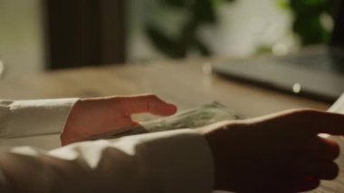 100 dolarlık banknotu ahşap bir masanın üzerinden kaydıran bir el, profesyonel bir ofis ortamının ışığı altında yakalanmış, bu da finansal bir işleme işaret ediyor.