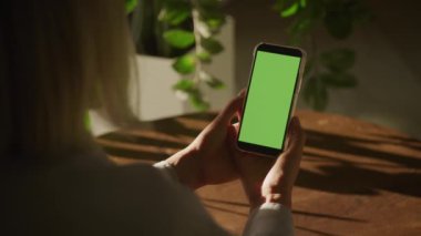 Doğal ışık ve kapalı alan bitkileriyle çevrili yeşil ekranlı akıllı telefonu tutan bir kadının elleri.