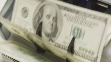 Yüz dolarlık banknotların yakın çekim görüntüsü bir para sayma makinesinde işleniyor. Görüntü, büyük miktarda para kullanmanın hareketini ve otomasyonunu yakalar.