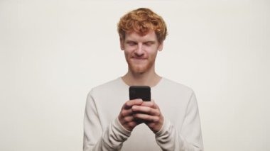 Kızıl saçlı bir adam akıllı bir telefon tutuyor ve beyaz arka planda dalgın dalgın bakıyor.