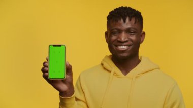 Sarı kapüşonlu genç adam sarı arka planda yeşil ekranlı akıllı telefonu gösteriyor.