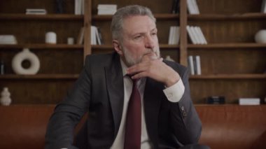 Düşünceli bir ifadeye sahip takım elbiseli, kitap rafı arka planında bir ofis ortamında oturan olgun bir işadamı.