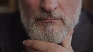 Olgun bir adamın sakalını, eli çenesinde düşünceli bir jest yaparak, yüz kıllarını ve dokusunu ön plana çıkararak yakın plan çekiyoruz.