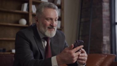 Akıllı telefon kullanırken gülümseyen takım elbiseli olgun bir işadamı, arka planda bir kitaplık ile bir ofis ortamında oturuyor.