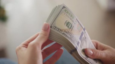 Yakın plan çekimde, iyi aydınlatılmış bir odada büyük bir deste Amerikan doları tutuyor ve dikkatle sayıyor.