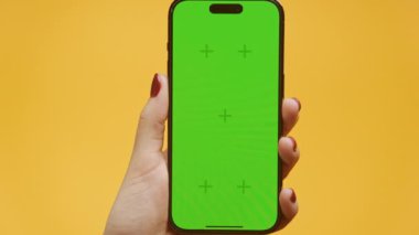 Kırmızı tırnaklı Zoom Out Hand sarı arka planda yeşil ekranlı akıllı telefonu tutuyor