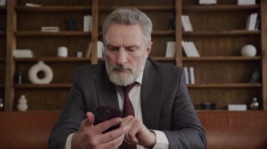 Takım elbiseli kıdemli işadamı, modern ofiste kütüphane geçmişi olan telefonları kontrol ediyor.