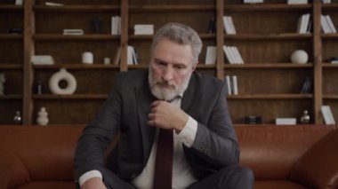 Takım elbiseli kıdemli işadamı, modern ofis ortamında oturuyor ve derin düşünüyor.