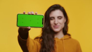 Sarı kapüşonlu yeşil ekranlı, sarı arka planda gülümseyen ve telefonu tutan kadın.