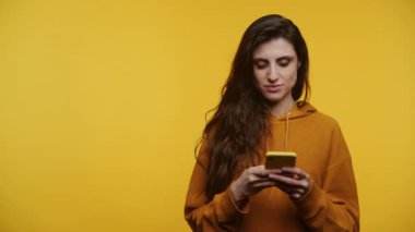 Sarı kapüşonlu gülümseyen kadın parlak sarı arka planda akıllı telefon kullanıyor.