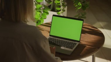 Yeşil bitkilerle güneşli bir odada yeşil ekranlı dizüstü bilgisayarda çalışan bir kadın.
