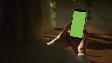 Elinde yeşil bitkilerle güneşli bir odada yeşil ekranlı akıllı bir telefon tutan kişi.
