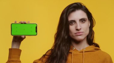 Sarı kapüşonlu kadın yeşil ekranlı akıllı telefonu sarı arka planda tutuyor, ciddi görünüyor, hoşnutsuzluğunu veya hoşnutsuzluğunu dile getiriyor.