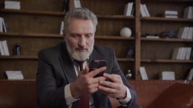 Takım elbiseli yaşlı bir adam telefonuyla video görüşmesi yapıyor, modern bir ofiste oturuyor.