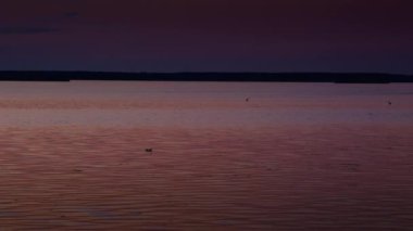 Sıcak turuncu ve mor renkler ile huzurlu bir gün batımı gölün sakin suları üzerinde yansıyor, kuşlar uzaklardan uçuyor.