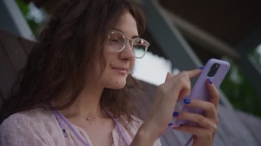 Kampüste akıllı telefon kullanan gözlüklü bir kadın, doğal arka planı olan ekrana odaklanıyor.