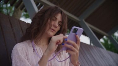 Kıvırcık saçlı bir kadın, açık havada tahta bir bankta otururken akıllı telefon kullanıyor.