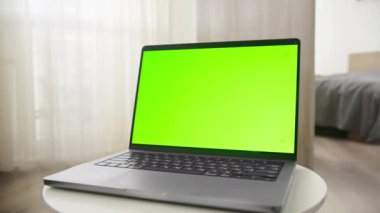 Düz perdeleri ve arka planında yatağı olan parlak bir odada, küçük bir masada yeşil ekranlı bir laptop.