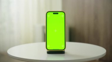 Parlak bir odada, küçük bir masada yeşil ekranlı akıllı bir telefon. Perdeleri ve ahşap bir arka planı var.