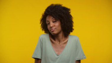 Kıvırcık saçlı ve günlük kıyafetli bir kadın sarı arka planda omuz silkiyor.