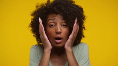 Kıvırcık saçlı bir kadın sarı arka planda yüzünü şokta tutuyor.