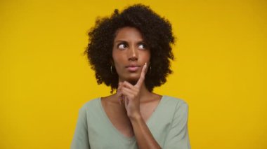 Kıvırcık saçlı kadın düşünceli görünüyor ve sarı arka planda parmağını çenesine koyuyor.