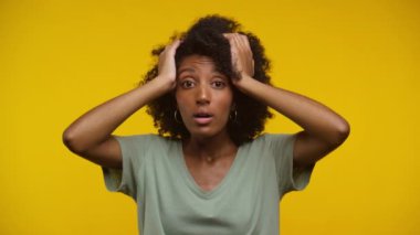Kıvırcık saçlı kadın şok olmuş gibi görünüyor, iki eliyle sarı arka planda başını tutuyor.