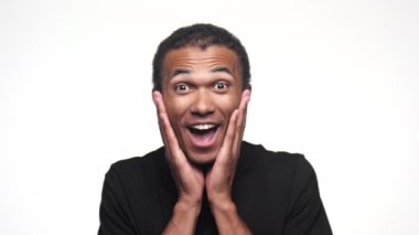 Siyah tişörtlü bir adam heyecanlı bir ifadeyle beyaz bir arka planda şaşkınlıkla yüzünü tutuyor.