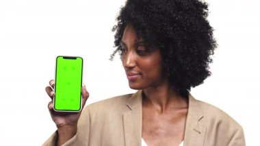 Kıvırcık saçlı genç bir kadının portresi. Elinde yeşil ekranı gösteren bir akıllı telefon tutarken gülümsüyor. Beyaz arka planda bej bir ceket giyiyor.