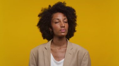 Kıvırcık saçlı, spor ceket giyen, sarı arka planda duran, ciddi görünen kendine güvenen genç bir kadın.