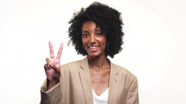 Kıvırcık saçlı genç bir kadın yüzünde bir gülümsemeyle barış işareti yapıyor, beyaz arka planda soyutlanmış.