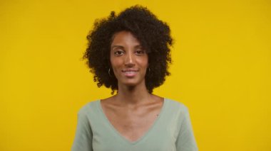 Doğal kıvırcık saçlı genç bir kadının yakın plan portresi, nazikçe gülümsüyor ve doğrudan kameraya bakıyor, parlak sarı bir arka plana karşı onay jestiyle başını sallıyor.